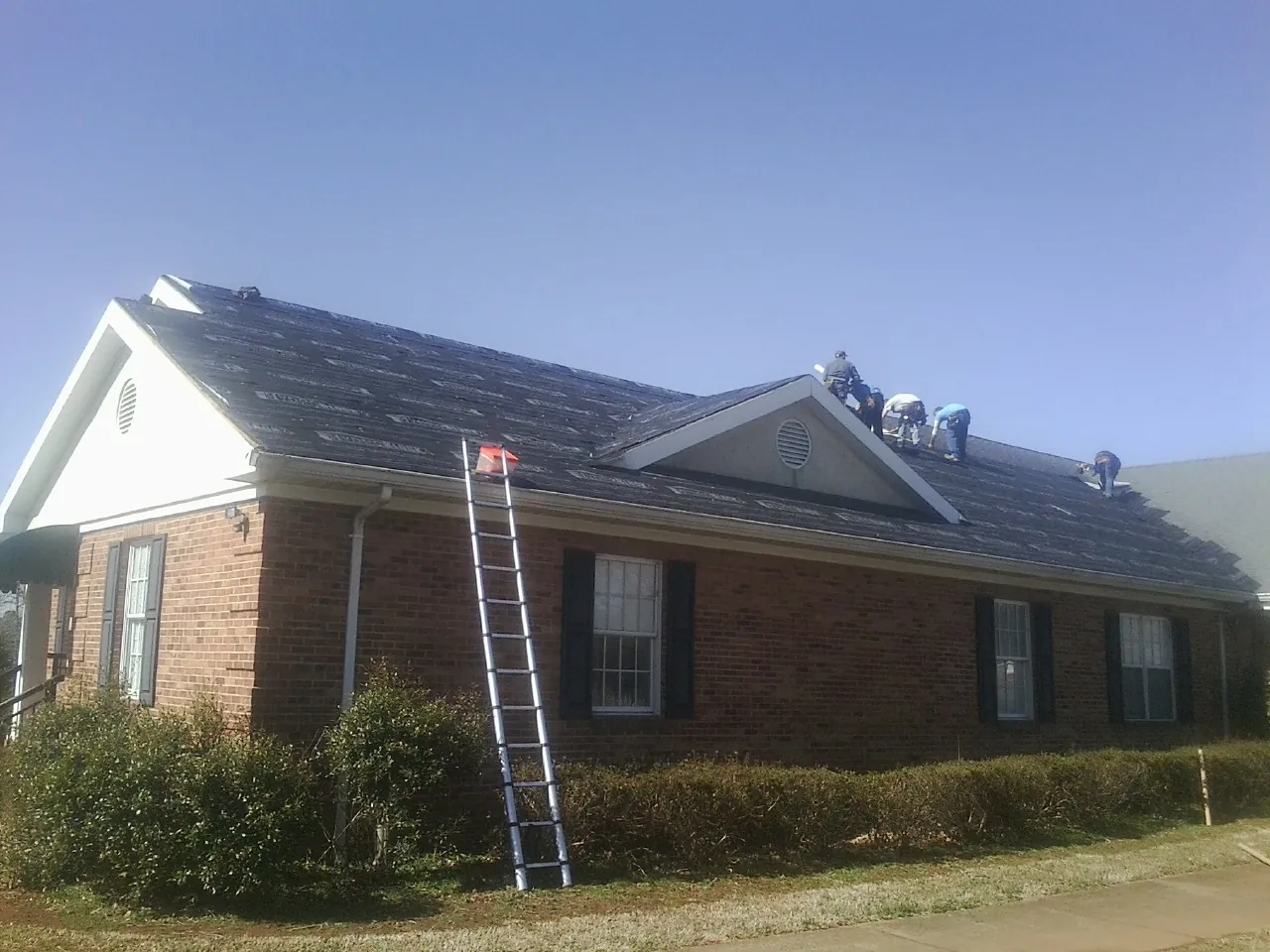 Crew tearing shingles off a brick ranch roof with a ladder set — Emergency Roof Repair work by our crew in Van Vleck