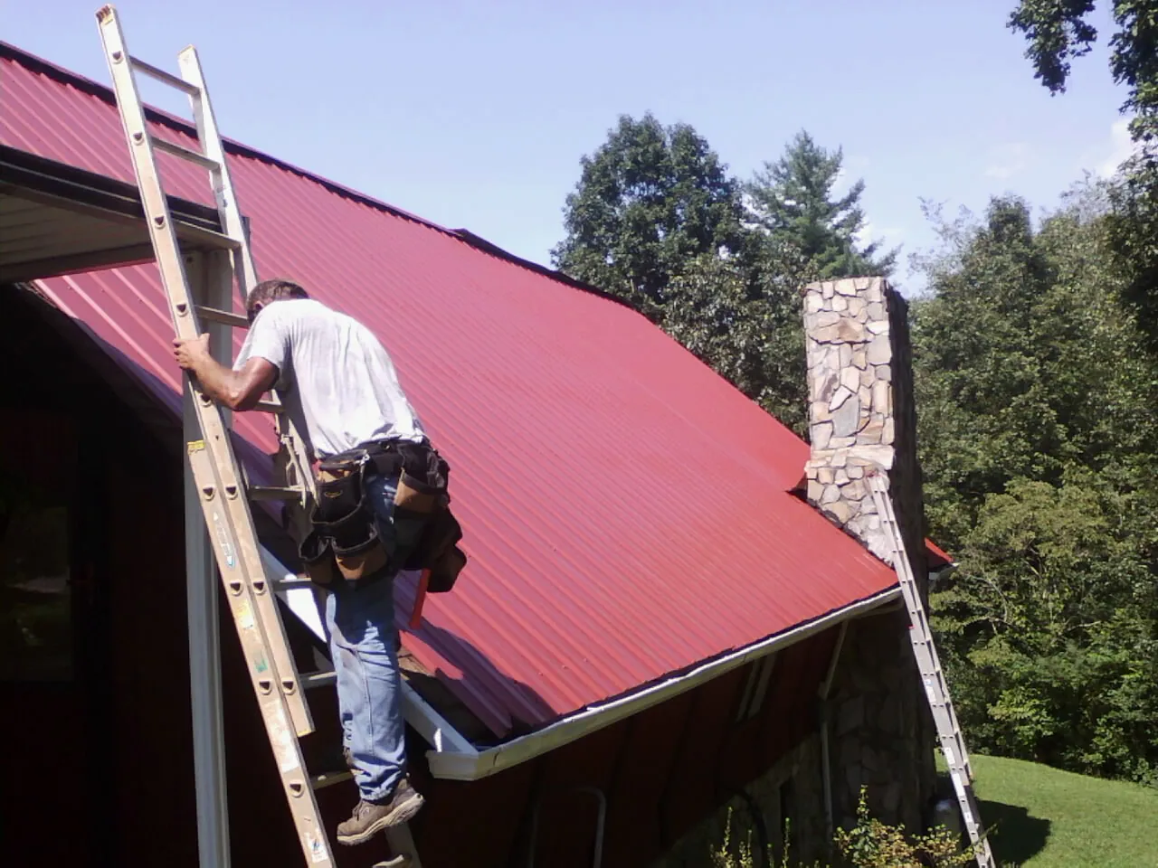 Roofer on a ladder beside a red metal roof and stone chimney — Soffit & Fascia Repair by Roofing Brothers in Van Vleck